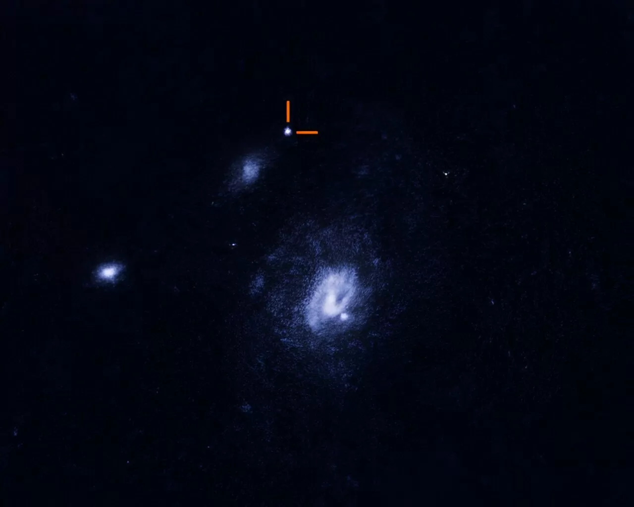 This captivating Hubble snapshot unveils a trio of galaxies, nestled amidst the serene black canvas of the cosmos. Dominating the frame is a grand spiral galaxy, adorned in hues of white and blue, stationed at the heart of the image. To its left, two smaller celestial islands of whitish hue make their presence known. Yet, it's the peculiar white fleck near the image's summit, underscored by a pair of intersecting orange lines, that steals the spotlight. This glow emanates from a mystifying exploded entity, unrelated to any of the galaxies in view, a cosmic enigma awaiting unravelling. | Credit: NASA, ESA, STScI, A. Chrimes (Radboud University)